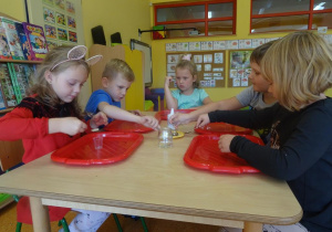 Dzieci siedzą przy stoliku i za pomocą łyżeczek przesypują drożdże z talerzyka na środku stolika do swoich małych plastikowych pojemników.